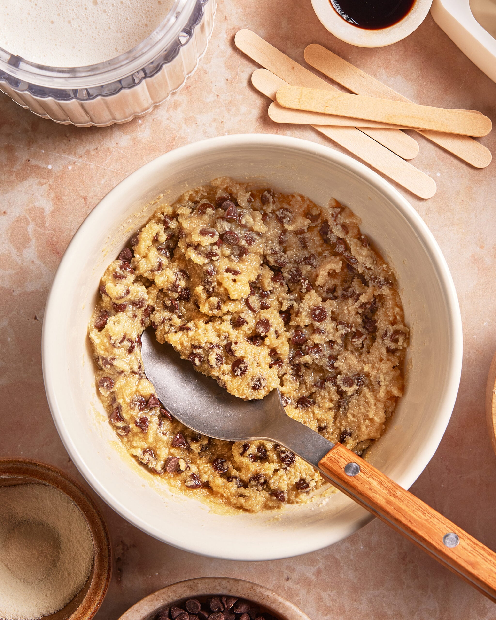 A mixing bowl filled with chocolate chip cookie dough and a spoon. Surrounding the bowl are bowls of ingredients, a stack of wooden sticks, and a cup of liquid on a marble countertop.