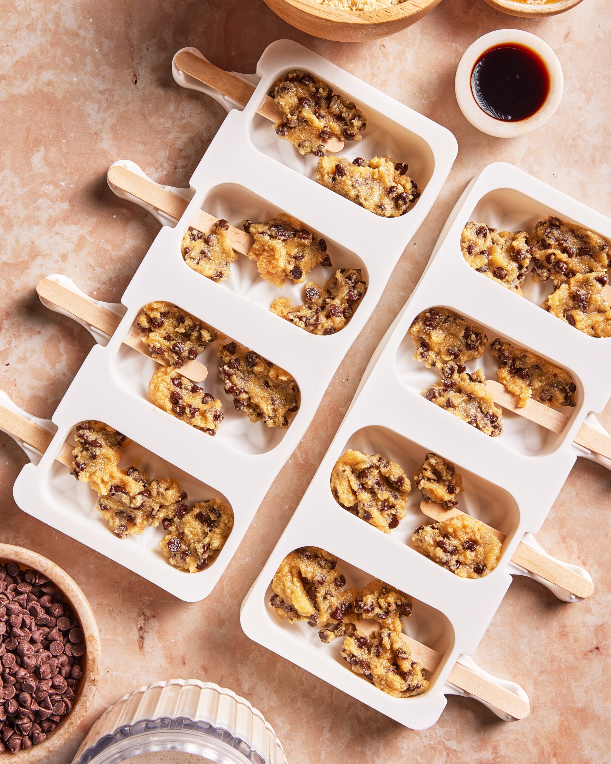 Two white silicone molds with wooden sticks are filled with unbaked chocolate chip cookie dough. Surrounding the molds are chocolate chips, brown sugar, and a small bowl of vanilla extract on a tan surface.