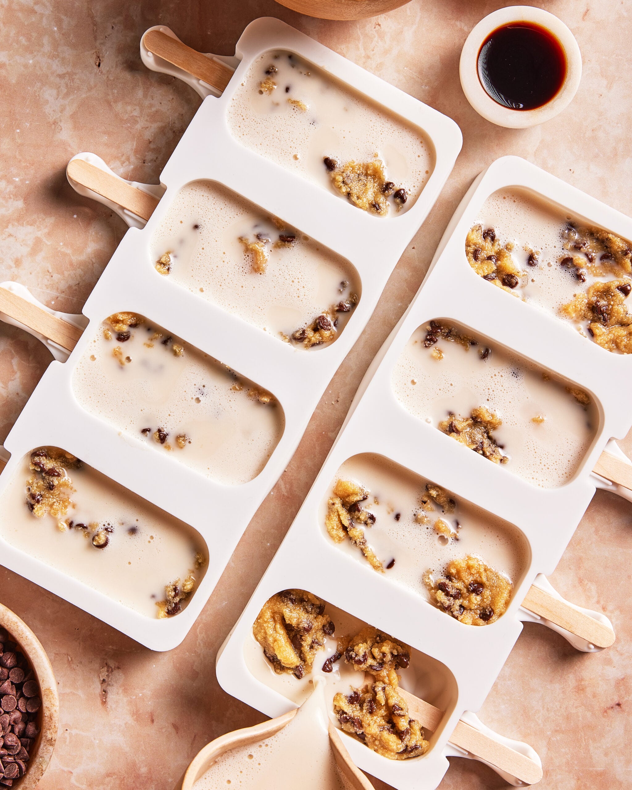 Two white silicone molds filled with a creamy mixture and cookie dough chunks on a tan surface, ready to freeze as popsicles. A bowl of chocolate chips and small bowls of sauce are nearby.