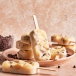 A plate with several creamy popsicles containing chocolate chips, one popsicle standing upright, with a bowl of chocolate chips in the background on a marble surface.