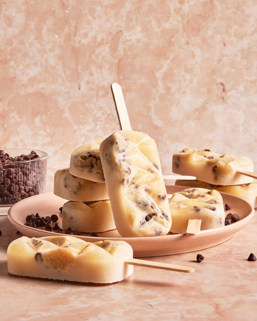 A plate with several creamy popsicles containing chocolate chips, one popsicle standing upright, with a bowl of chocolate chips in the background on a marble surface.