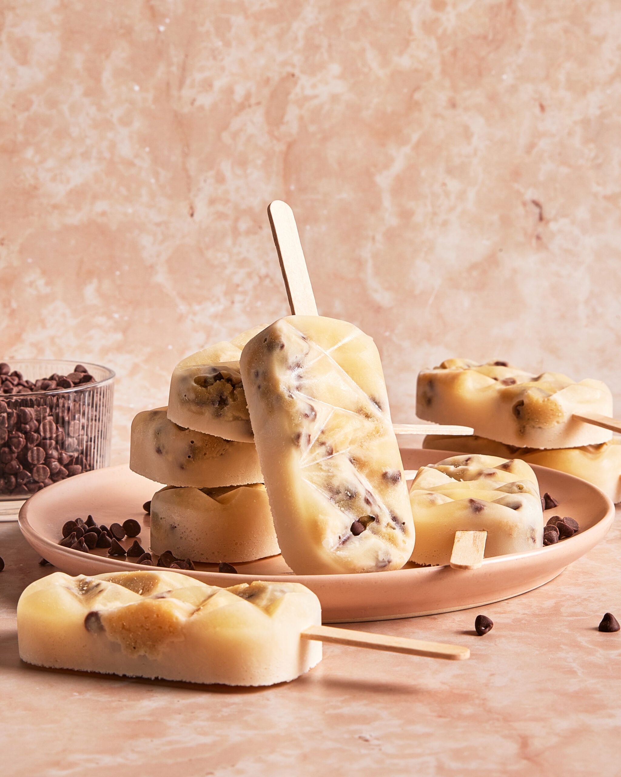 A plate with several creamy popsicles containing chocolate chips, one popsicle standing upright, with a bowl of chocolate chips in the background on a marble surface.