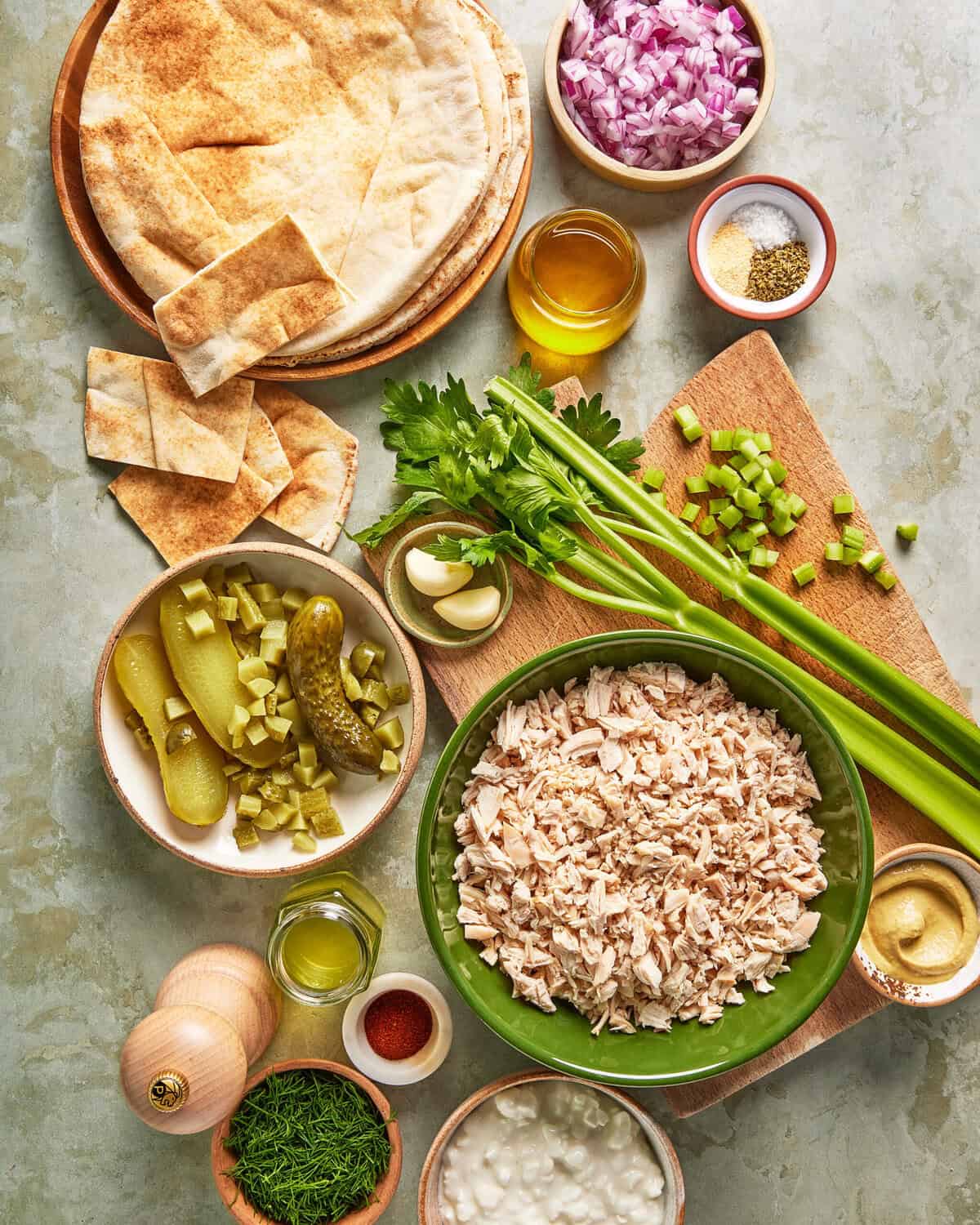Chopped chicken, celery, red onion, pickles, herbs, spices, pita bread, cottage cheese, oil, and mustard arranged on a table, with ingredients in bowls and on a wooden board, viewed from above.