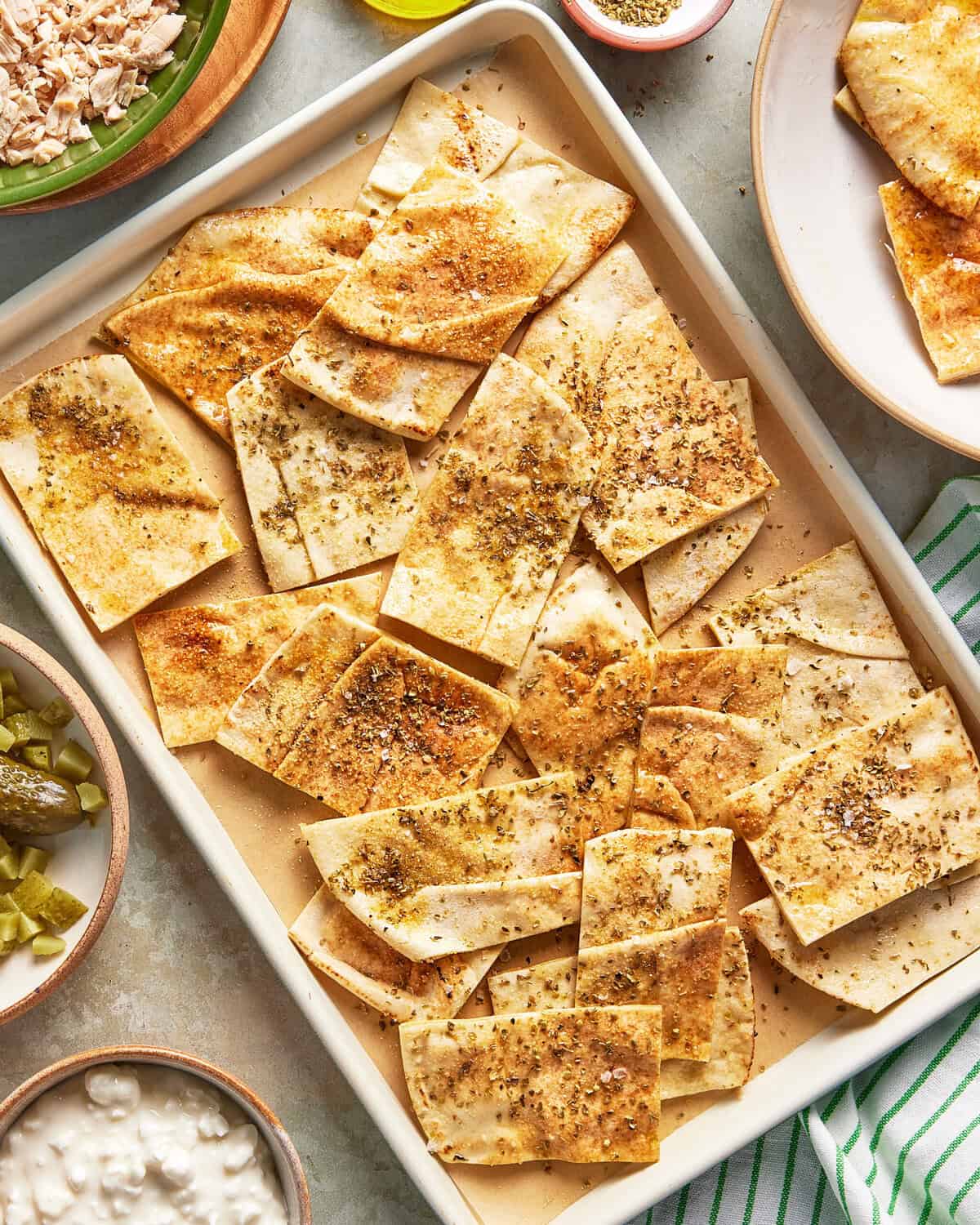 A tray of baked flatbread crackers cut into rectangles, topped with assorted seasonings, surrounded by small bowls of dips, pickles, and a plate with more flatbread on a light surface.