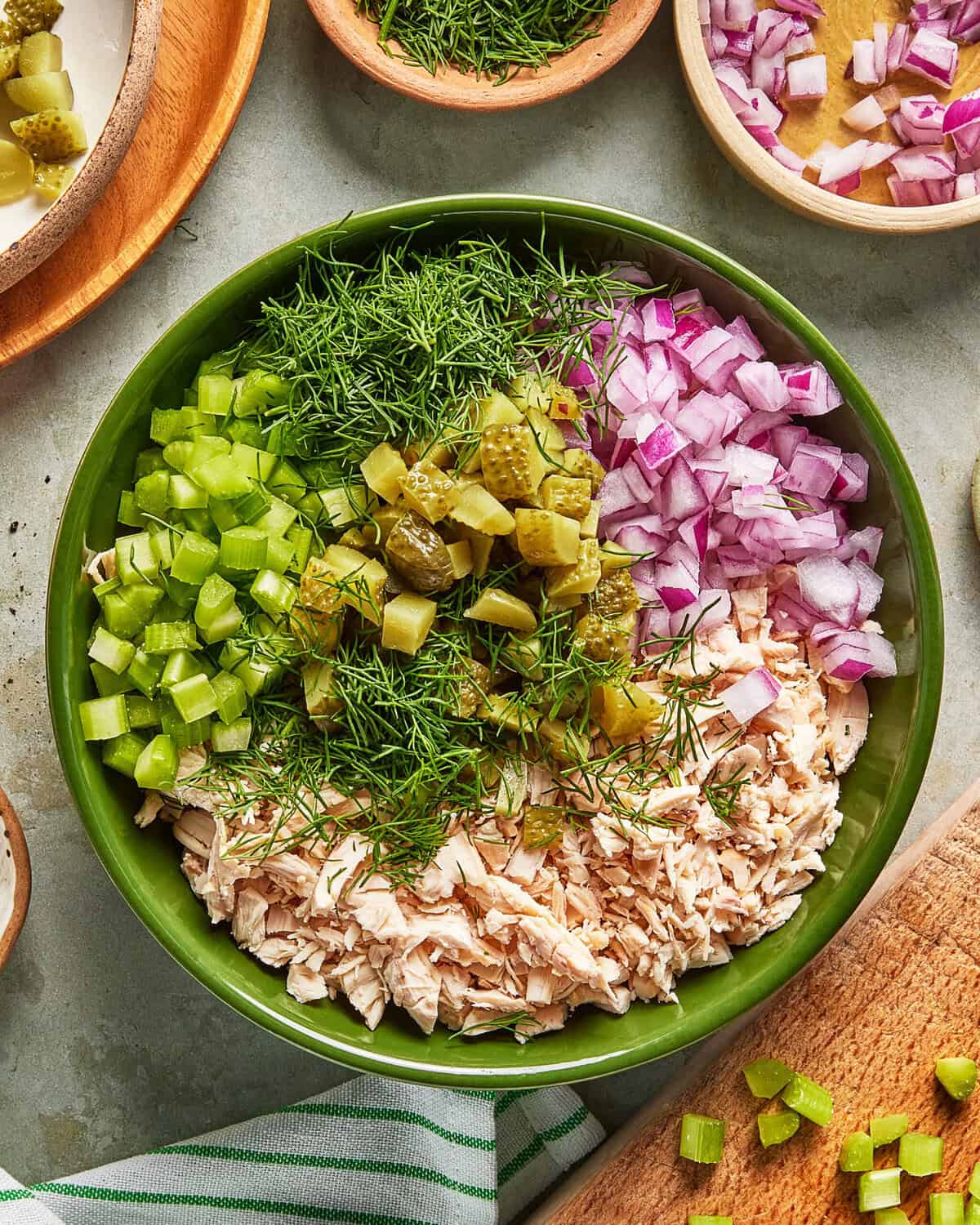 A green bowl filled with chopped celery, red onion, dill, pickles, and shredded chicken, ready to be mixed. Other small bowls and cutting board with more ingredients surround it.