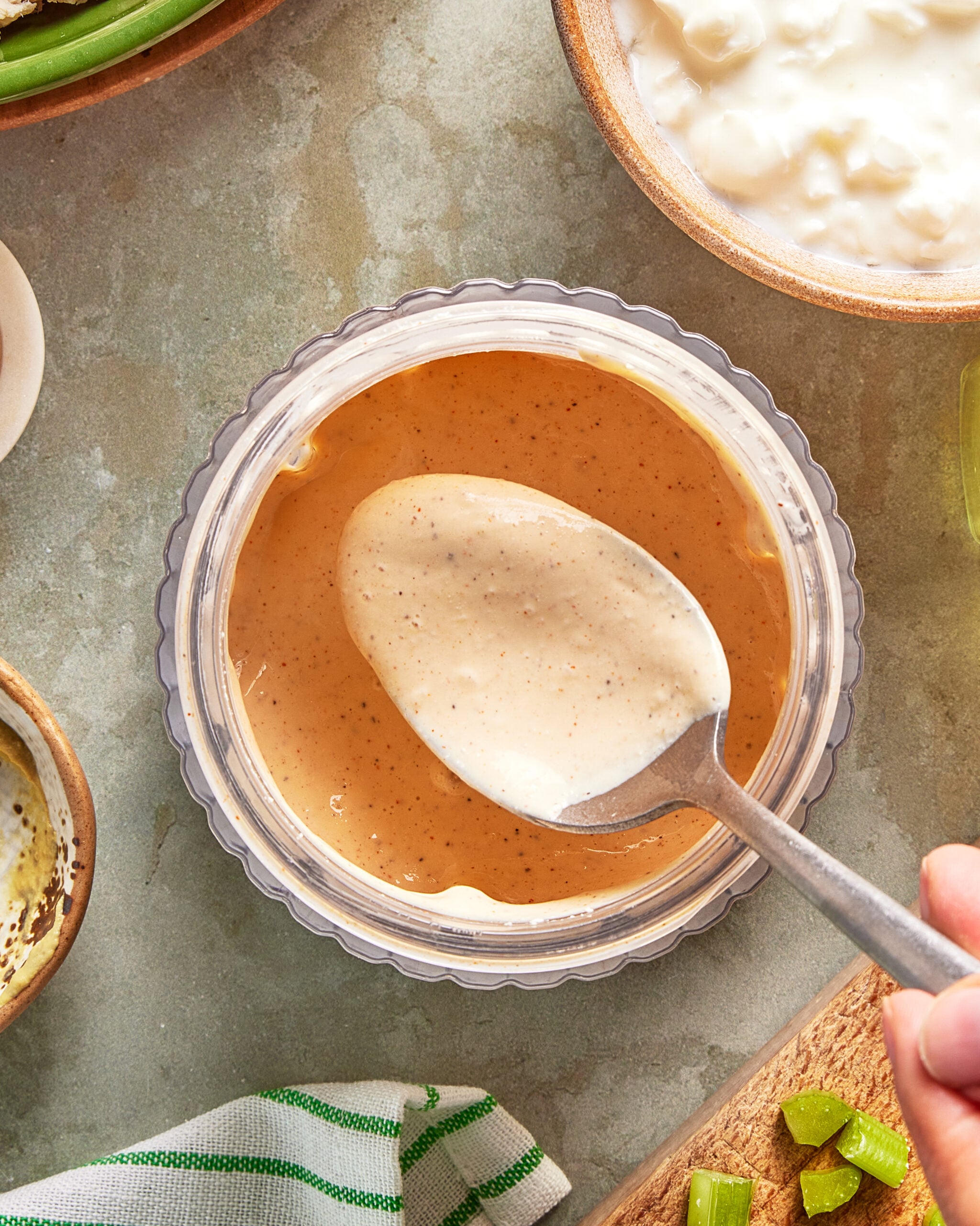A hand holding a spoon scoops up some creamy, light brown dipping sauce from a small glass bowl. Other bowls with dips and green celery pieces are nearby on a neutral-colored surface.