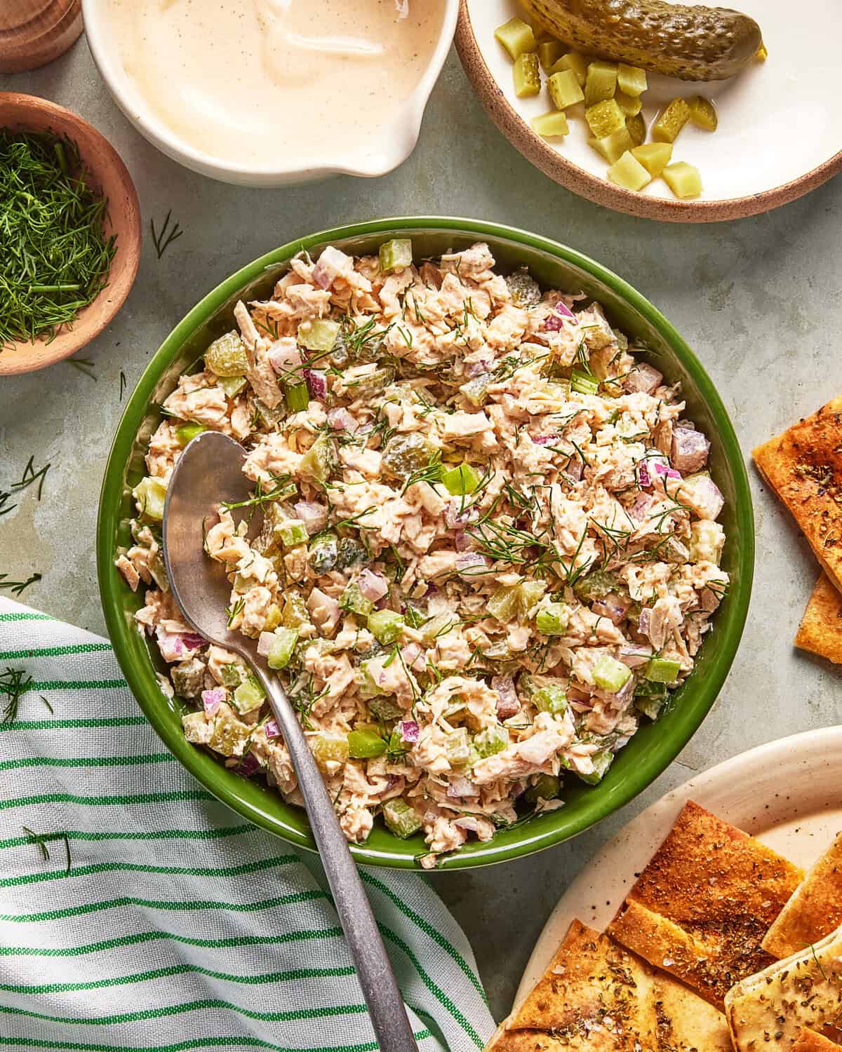 A bowl of chicken salad with celery, pickles, and red onion, garnished with dill. Surrounded by pita chips, a pickle spear, chopped pickles, a creamy dressing, and a striped napkin. A spoon rests in the bowl.
