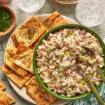 A green bowl of tuna salad with celery and red onions is surrounded by flatbread crackers, ice water, pickles, and fresh dill on a light-colored table.