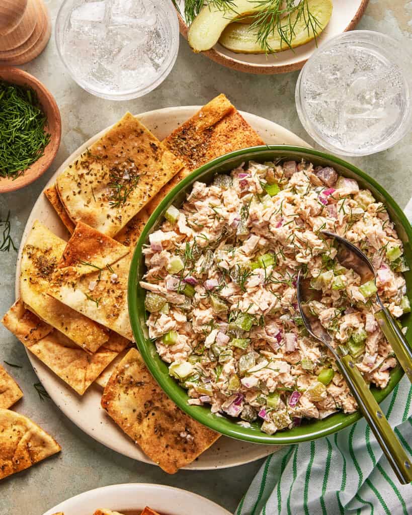 A green bowl of tuna salad with celery and red onions is surrounded by flatbread crackers, ice water, pickles, and fresh dill on a light-colored table.