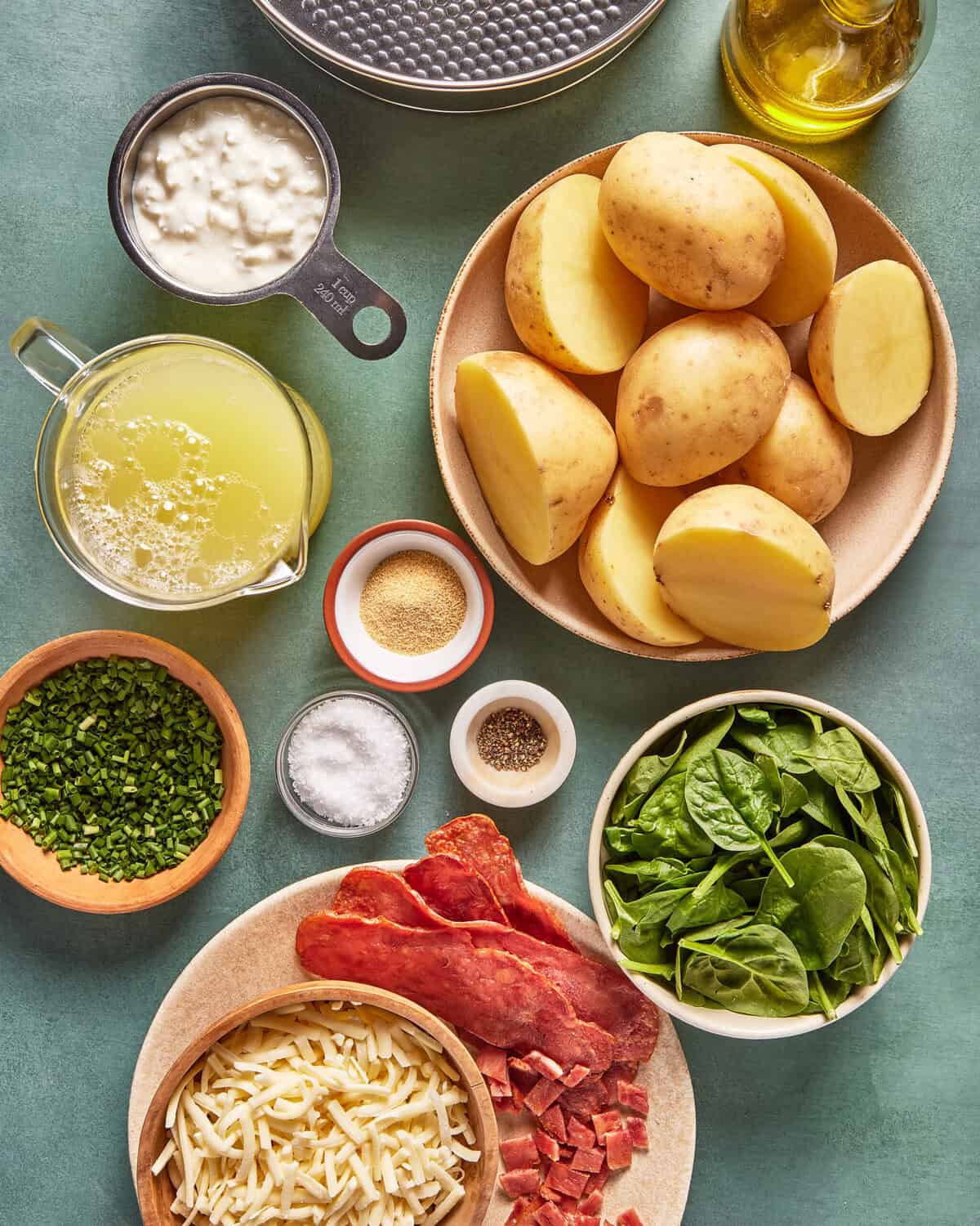 Sliced potatoes, fresh spinach, cottage cheese, shredded cheese, cooked bacon, chives, broth, oil, salt, pepper, and seasoning are arranged neatly on a table with a springform pan above.