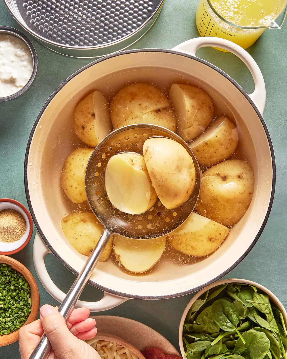 A hand holds a slotted spoon lifting a boiled potato wedge from a pot filled with other potato pieces, with bowls of spinach, chives, cheese, and other ingredients nearby on a green surface.