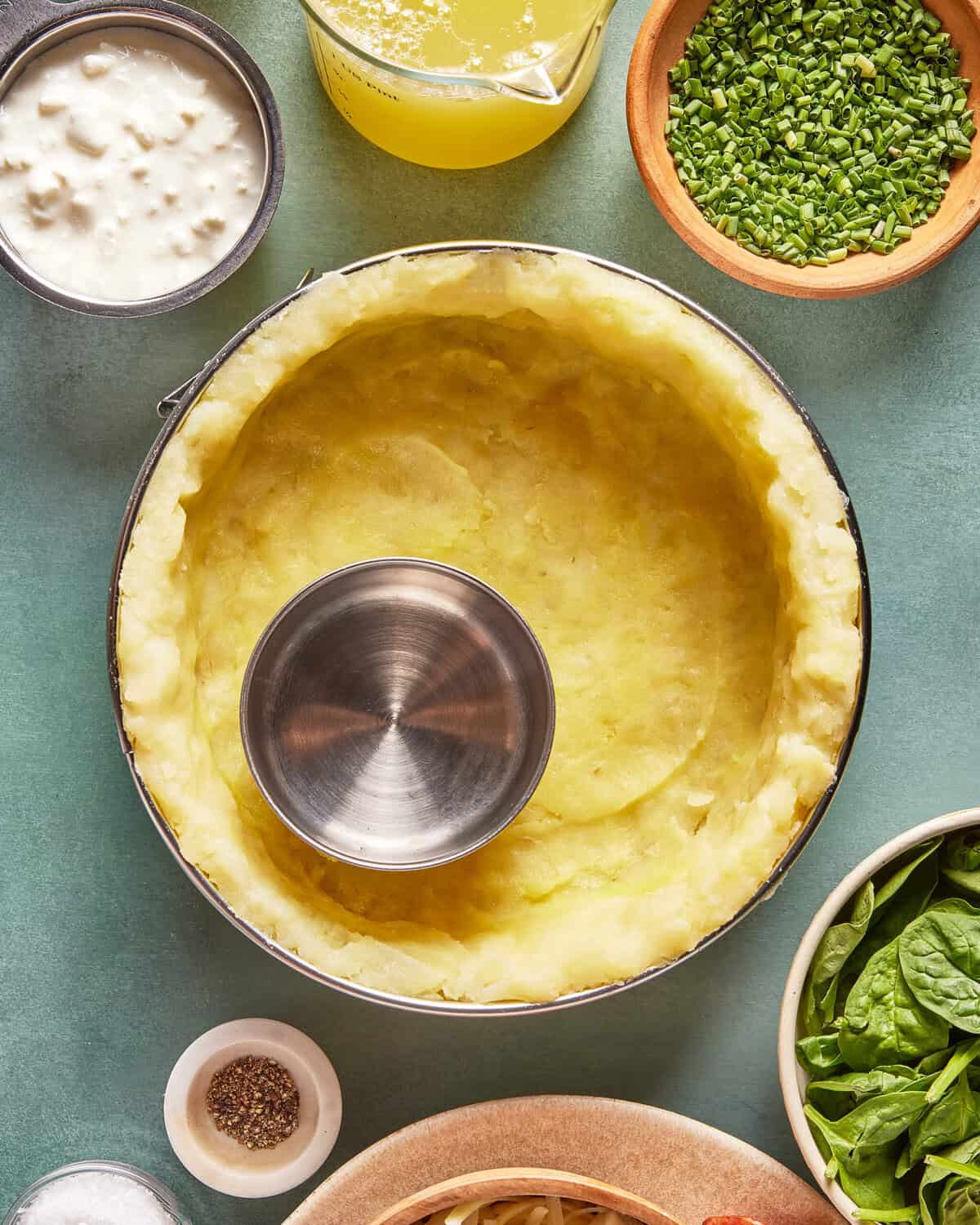 A metal ring presses into mashed potatoes in a round pan, surrounded by bowls of cottage cheese, chopped chives, spinach, pepper, and broth on a green surface.