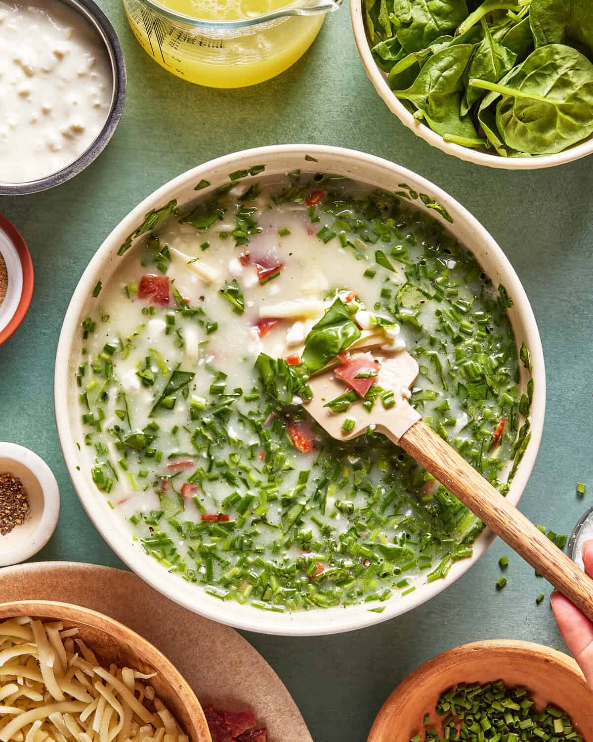 A bowl of creamy soup with chopped herbs, bits of bacon, and a spatula, surrounded by bowls of spinach, cheese, chives, and seasonings on a green tabletop.