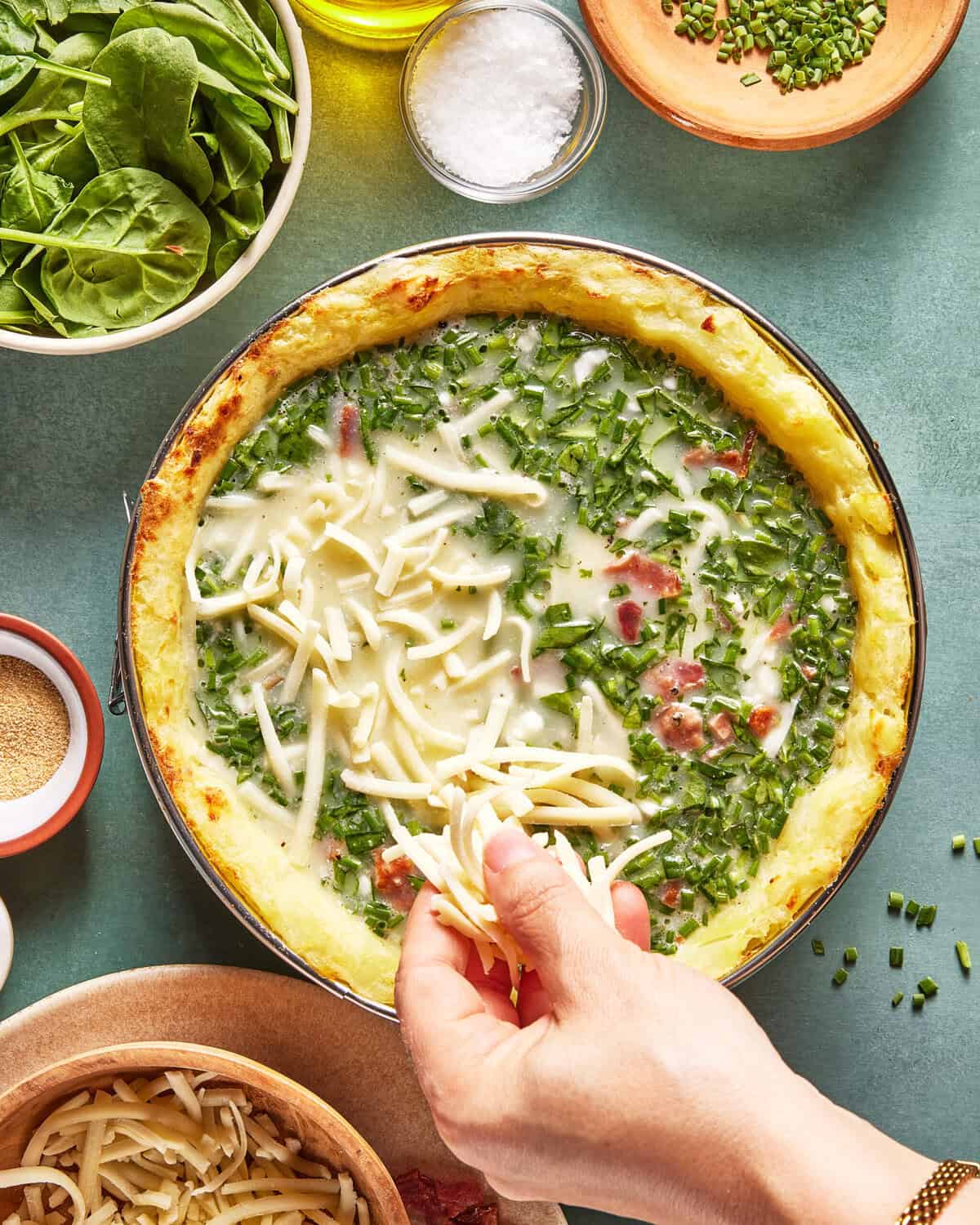 A hand sprinkles shredded cheese onto a quiche filled with greens and cream, surrounded by bowls of fresh spinach, chives, salt, and seasonings on a green surface.