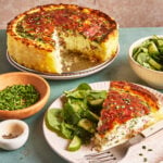 A slice of savory pie with a golden crust is served with fresh spinach and cucumber salad. The whole pie, partially sliced, sits nearby, along with bowls of chopped chives, salt, and pepper on a green surface.