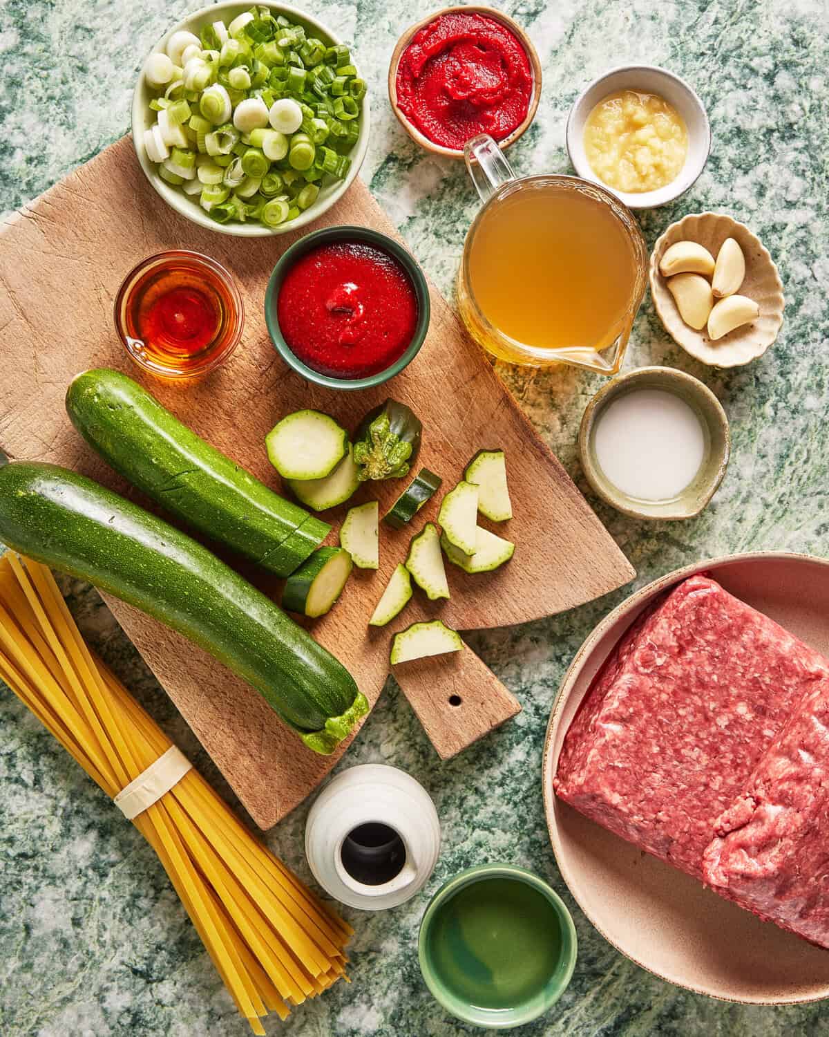 Ingredients on a counter: ground meat, zucchini, dry pasta, tomato paste, tomato sauce, broth, scallions, garlic, oil, cream, vinegar, and a small bowl of sliced green onions. All are set on a green marble surface.