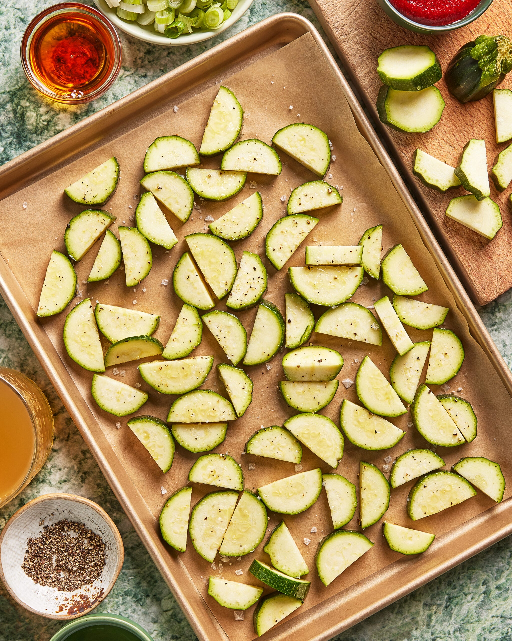 Sliced zucchini pieces spread on a parchment-lined baking sheet, sprinkled with salt and pepper. Various bowls and ingredients are visible around the tray on a green countertop.