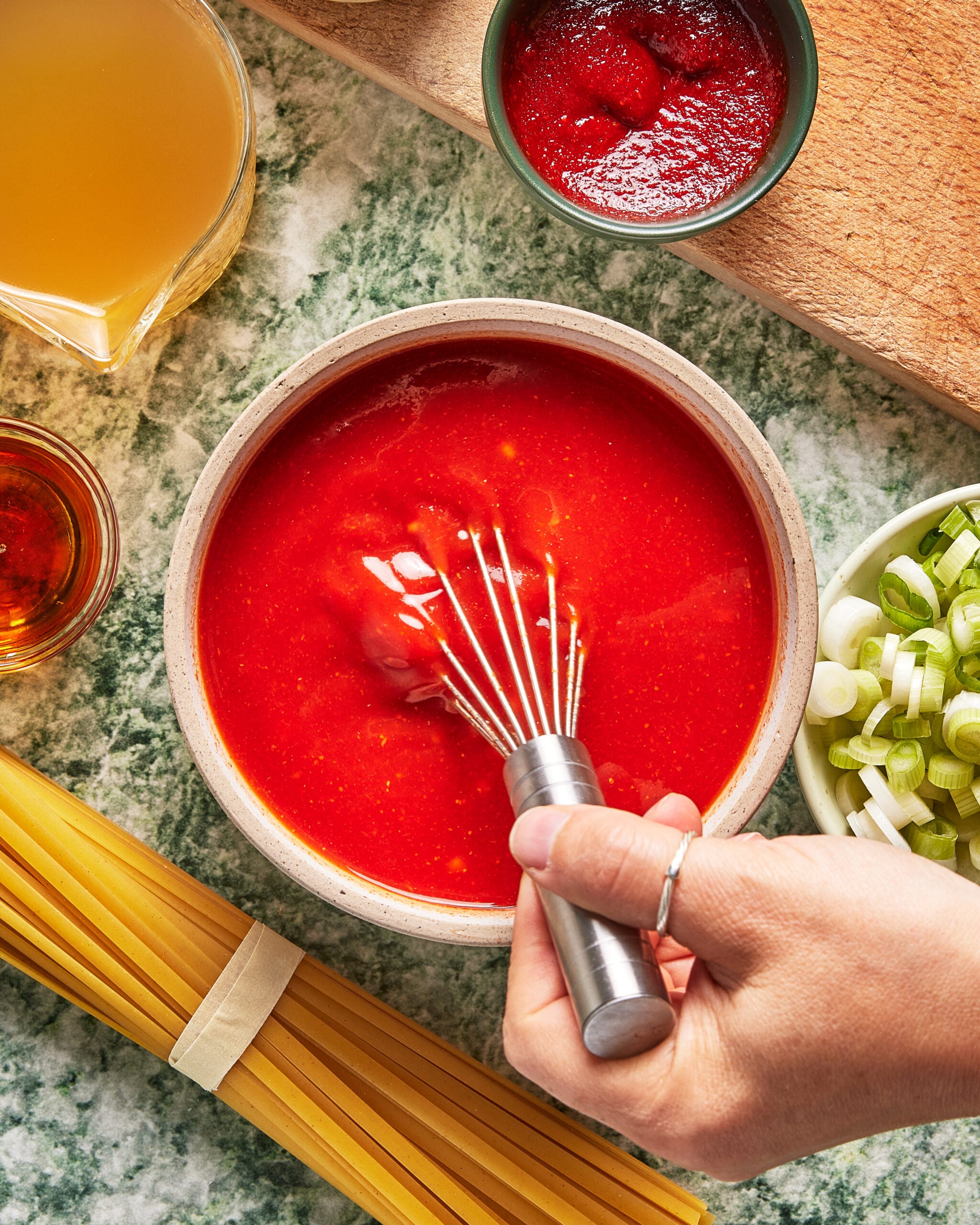 A hand whisks tomato sauce in a bowl, surrounded by uncooked spaghetti, chopped green onions, tomato paste, broth, and a small glass of liquid on a green marble countertop.