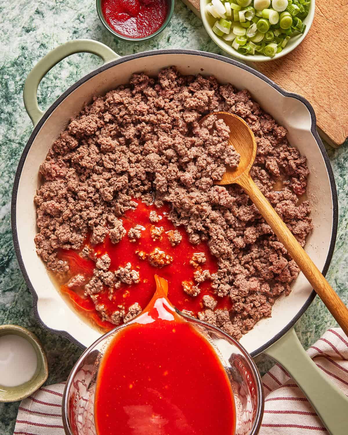 Ground beef cooking in a large skillet with tomato sauce being poured in, a wooden spoon inside, chopped green onions and tomato paste in bowls nearby, on a green countertop with a striped towel.