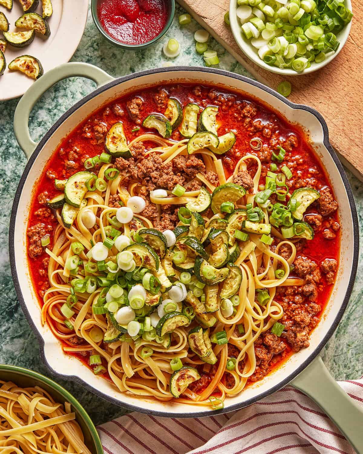 A skillet filled with linguine pasta, ground meat in tomato sauce, sautéed zucchini, and topped with chopped green onions. Surrounding the skillet are bowls of pasta, tomato paste, and a cutting board with green onions.
