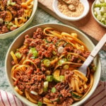 A bowl of noodles topped with minced meat, sliced zucchini, chopped green onions, and sauce. A fork is twirled in the noodles. Nearby are bowls of sesame seeds, sliced green onions, and a striped cloth napkin.