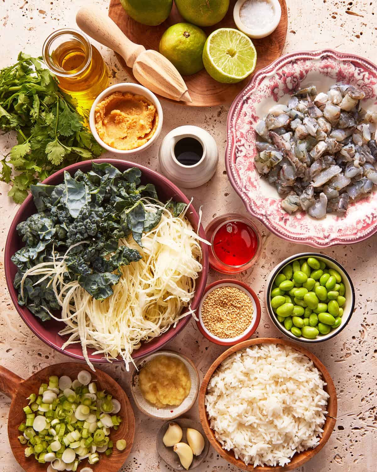 A top view of various ingredients in bowls and plates, including leafy greens, shredded cabbage, shrimp, white rice, edamame, limes, ginger, garlic, soy sauce, sesame seeds, oil, and seasonings on a textured surface.