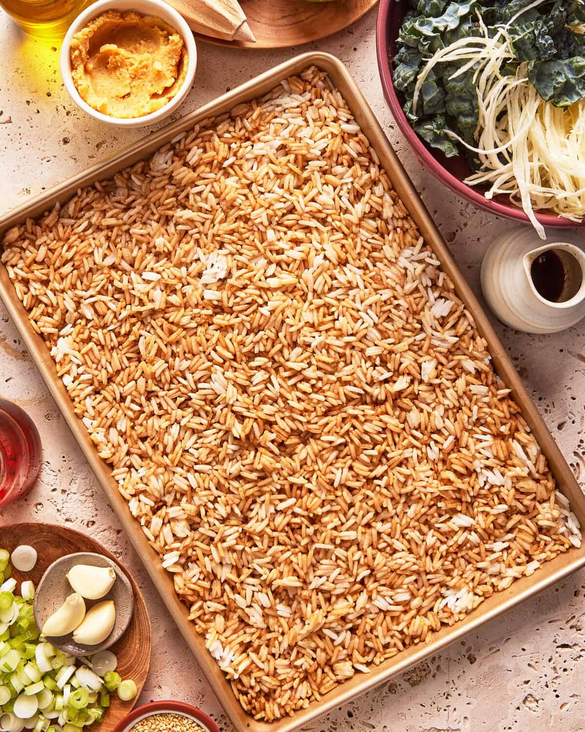 A sheet pan filled with uncooked orzo pasta is surrounded by small bowls of miso paste, torn kale, julienned vegetables, sliced green onions, garlic cloves, red liquid, oil, and seasonings on a textured surface.
