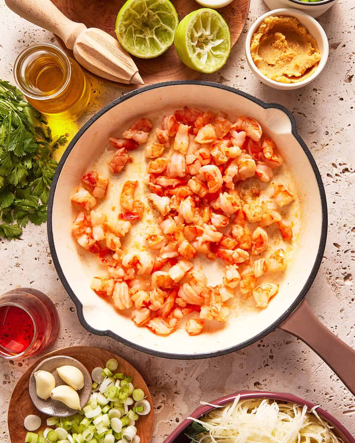 Chopped shrimp cooking in a white skillet surrounded by fresh ingredients, including lime halves, garlic cloves, green onions, herbs, oil, paste, and shredded vegetables on a beige countertop.