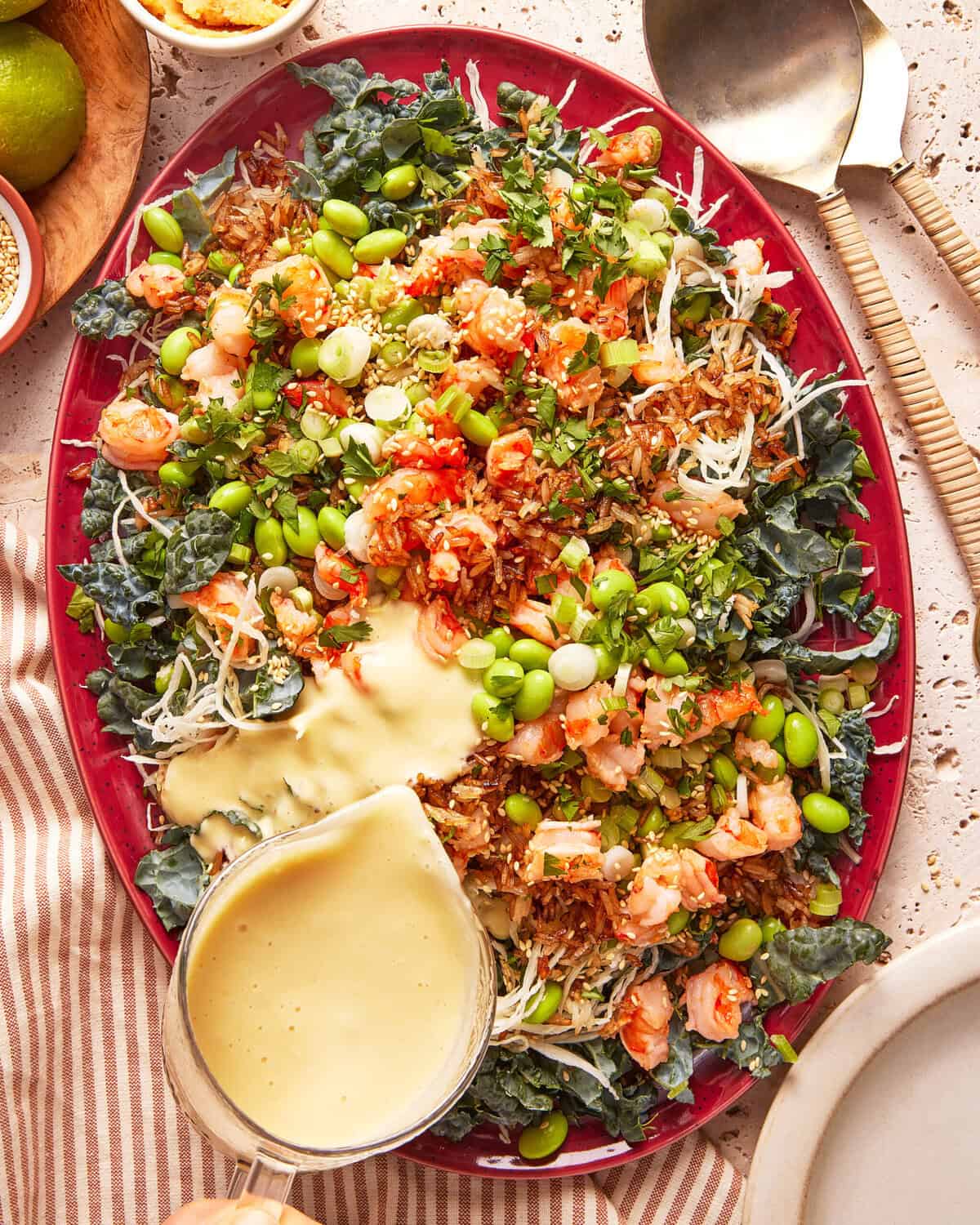 A vibrant salad on a red platter topped with shrimp, crispy onions, edamame, chopped greens, and fresh herbs, with a creamy yellow dressing being poured over one side. A large serving spoon rests nearby.