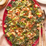 A vibrant salad on a red platter, featuring chopped dark leafy greens, edamame, shredded vegetables, shrimp, and sesame seeds, with serving utensils on the side and a striped cloth partially visible.