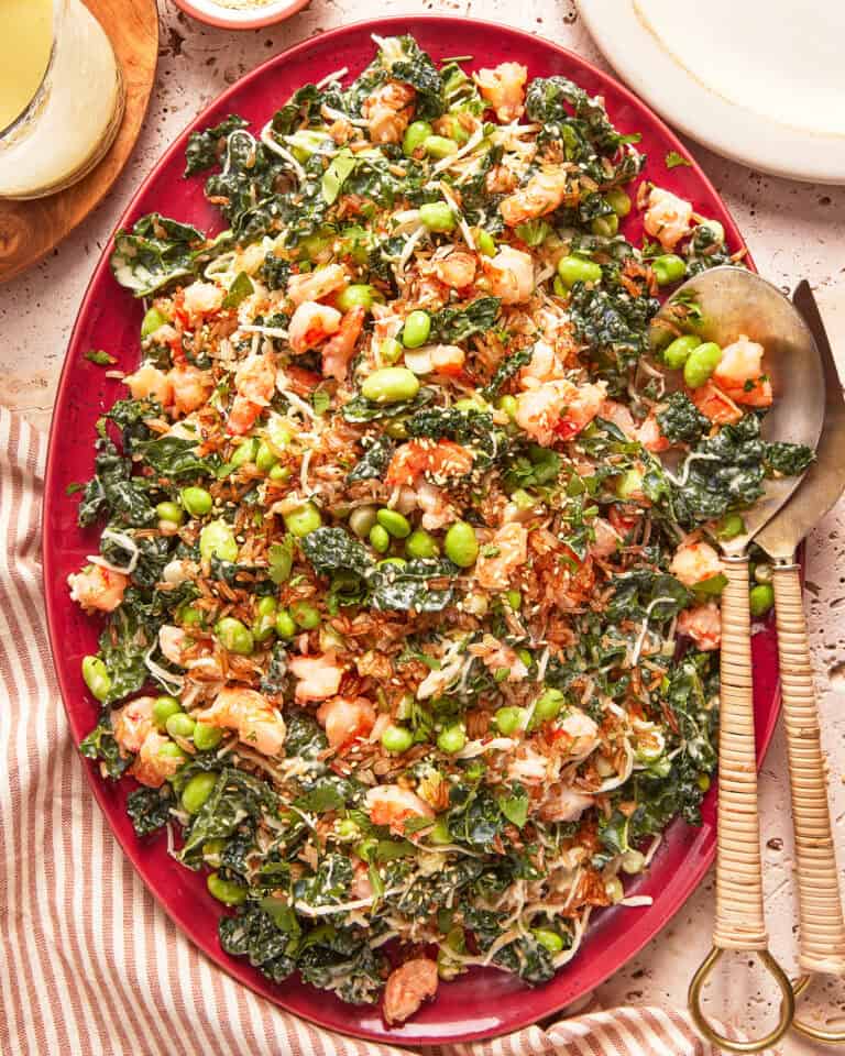 A vibrant salad on a red platter, featuring chopped dark leafy greens, edamame, shredded vegetables, shrimp, and sesame seeds, with serving utensils on the side and a striped cloth partially visible.