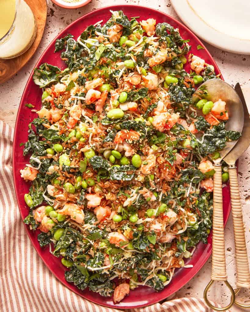 A vibrant salad on a red platter, featuring chopped dark leafy greens, edamame, shredded vegetables, shrimp, and sesame seeds, with serving utensils on the side and a striped cloth partially visible.