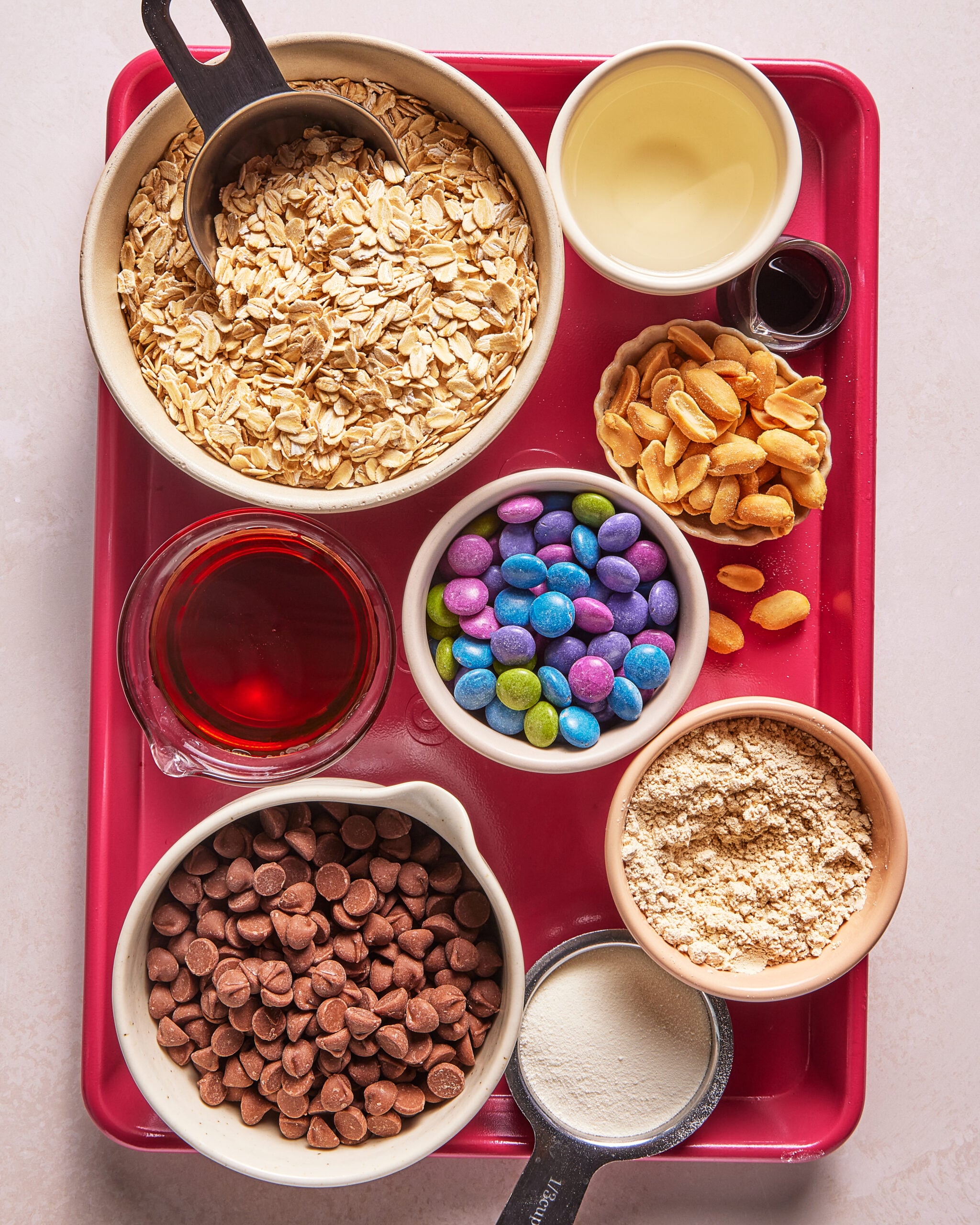 A red tray holding bowls of oats, maple syrup, oil, vanilla, peanuts, chocolate chips, colorful candy, protein powder, and peanut butter powder, seen from above.