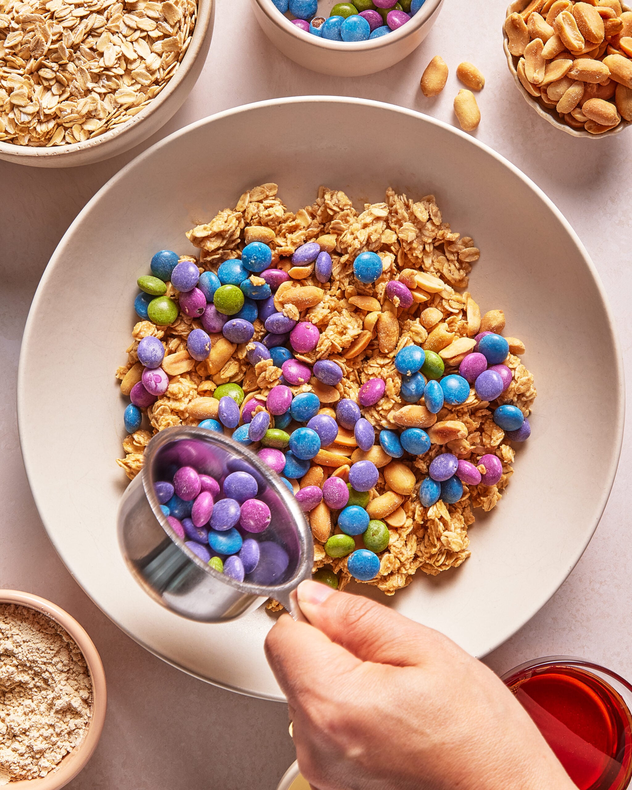 A hand adds colorful candy-coated chocolates to a bowl of granola, nuts, and oats, surrounded by bowls of oats, peanuts, flour, and a small cup of red liquid on a light surface.