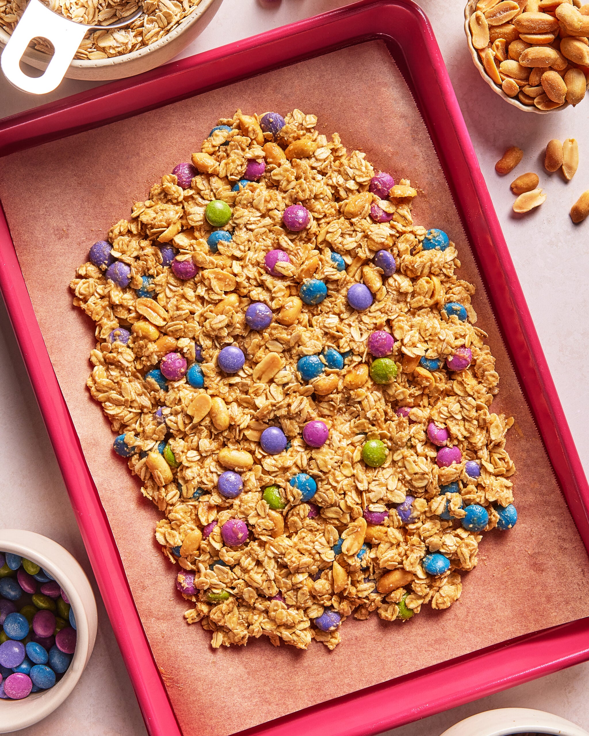 Granola mixture with colorful candy-coated chocolates shaped like an Easter egg on a baking sheet, surrounded by bowls of oats, peanuts, and candies.