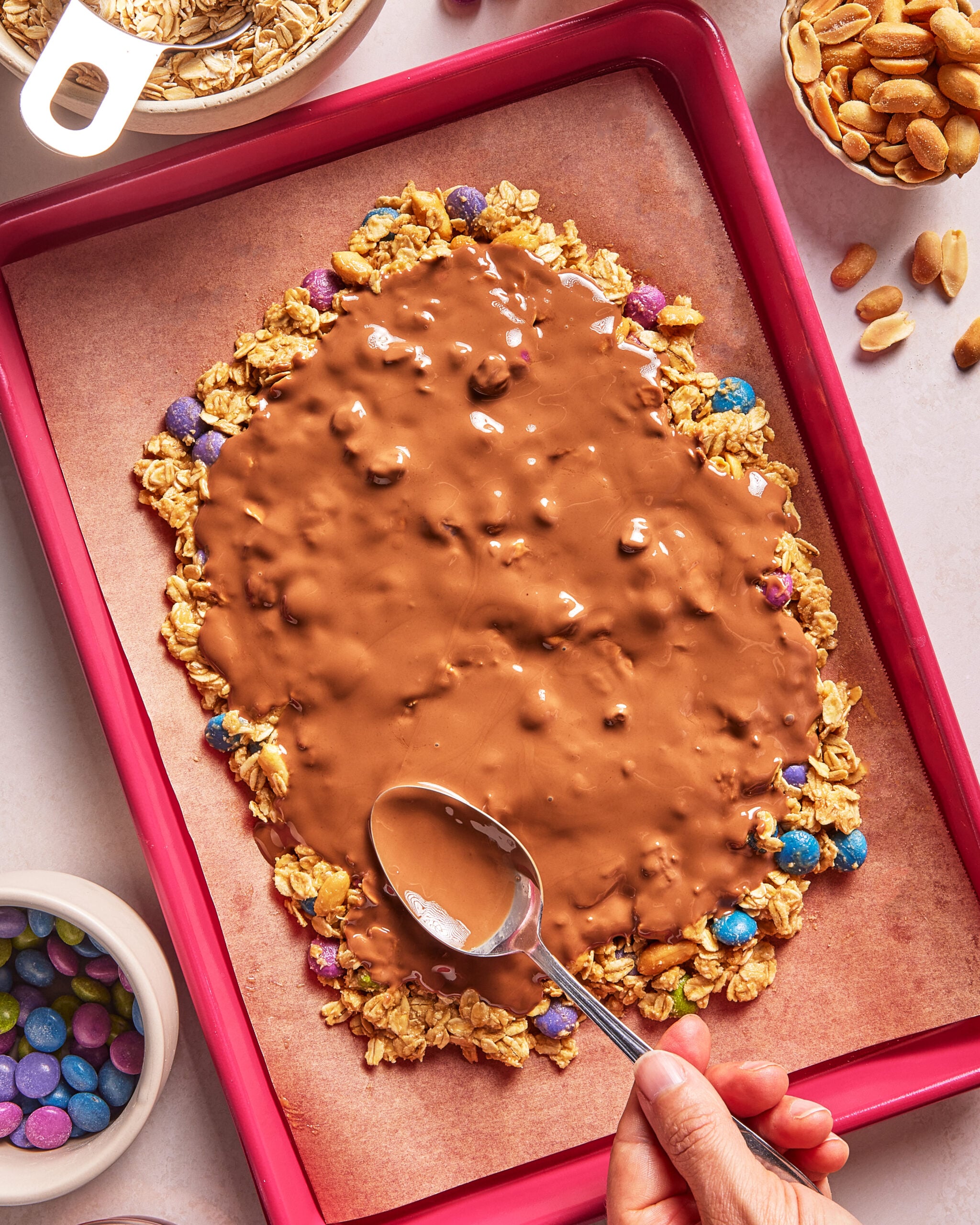 A hand spreads melted chocolate over a granola and candy layer on a baking sheet lined with parchment paper. Bowls of oats, peanuts, and colorful candy are nearby.