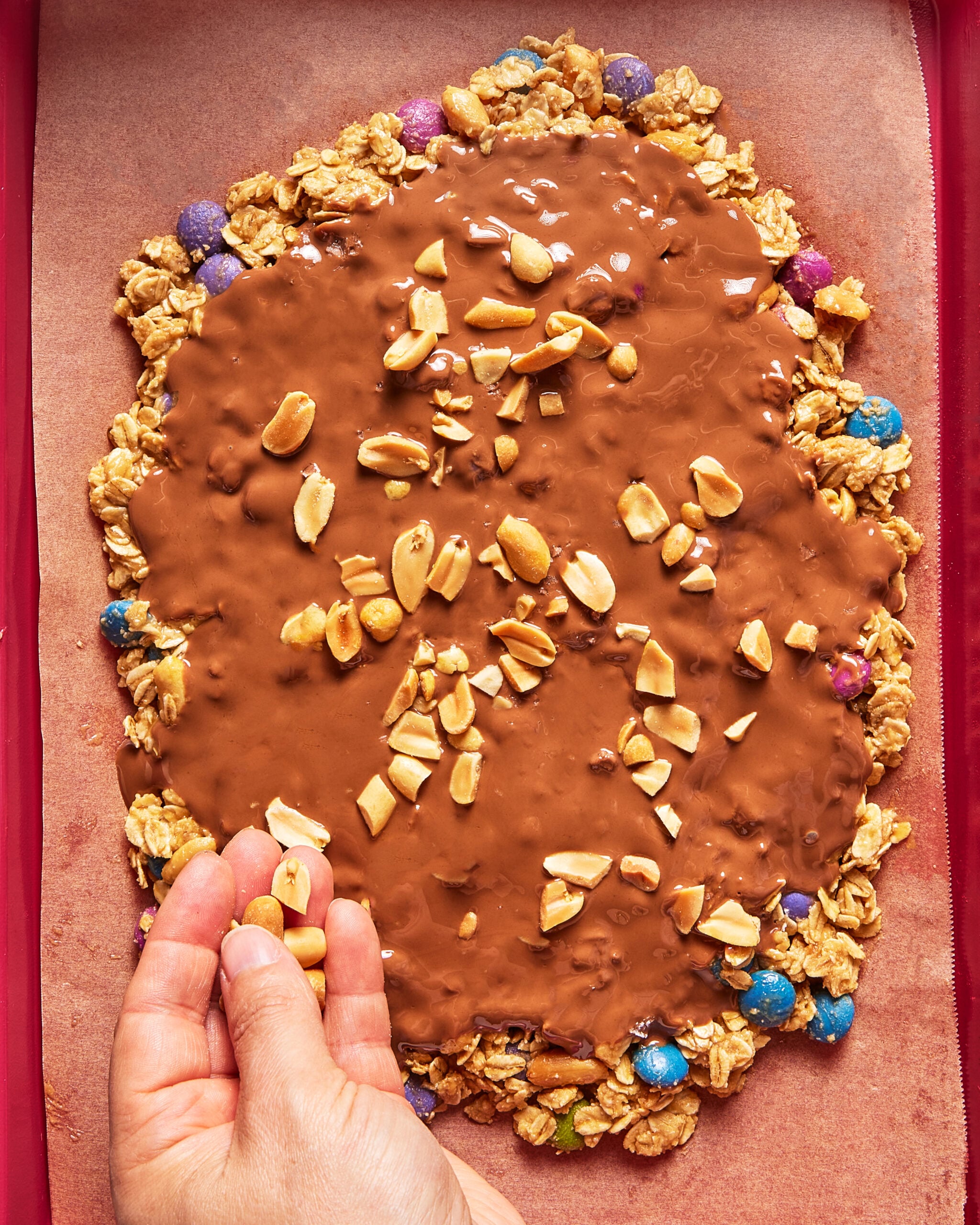 A hand sprinkles peanuts onto a granola mixture topped with melted chocolate and colorful candies, all on a sheet of parchment paper.