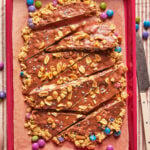 Granola bars topped with chocolate, peanuts, and sea salt, arranged on parchment paper in a red tray. Colorful candy-coated chocolates surround the bars.