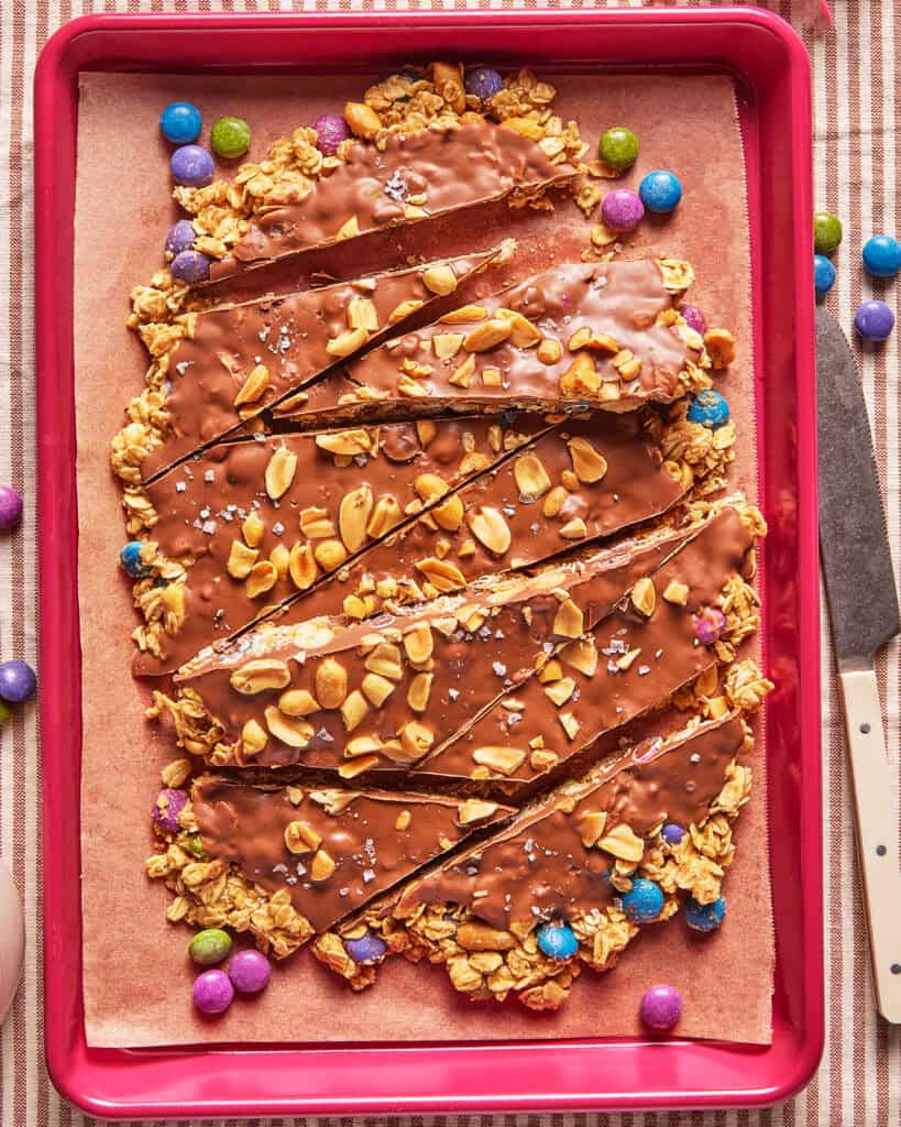 Granola bars topped with chocolate, peanuts, and sea salt, arranged on parchment paper in a red tray. Colorful candy-coated chocolates surround the bars.