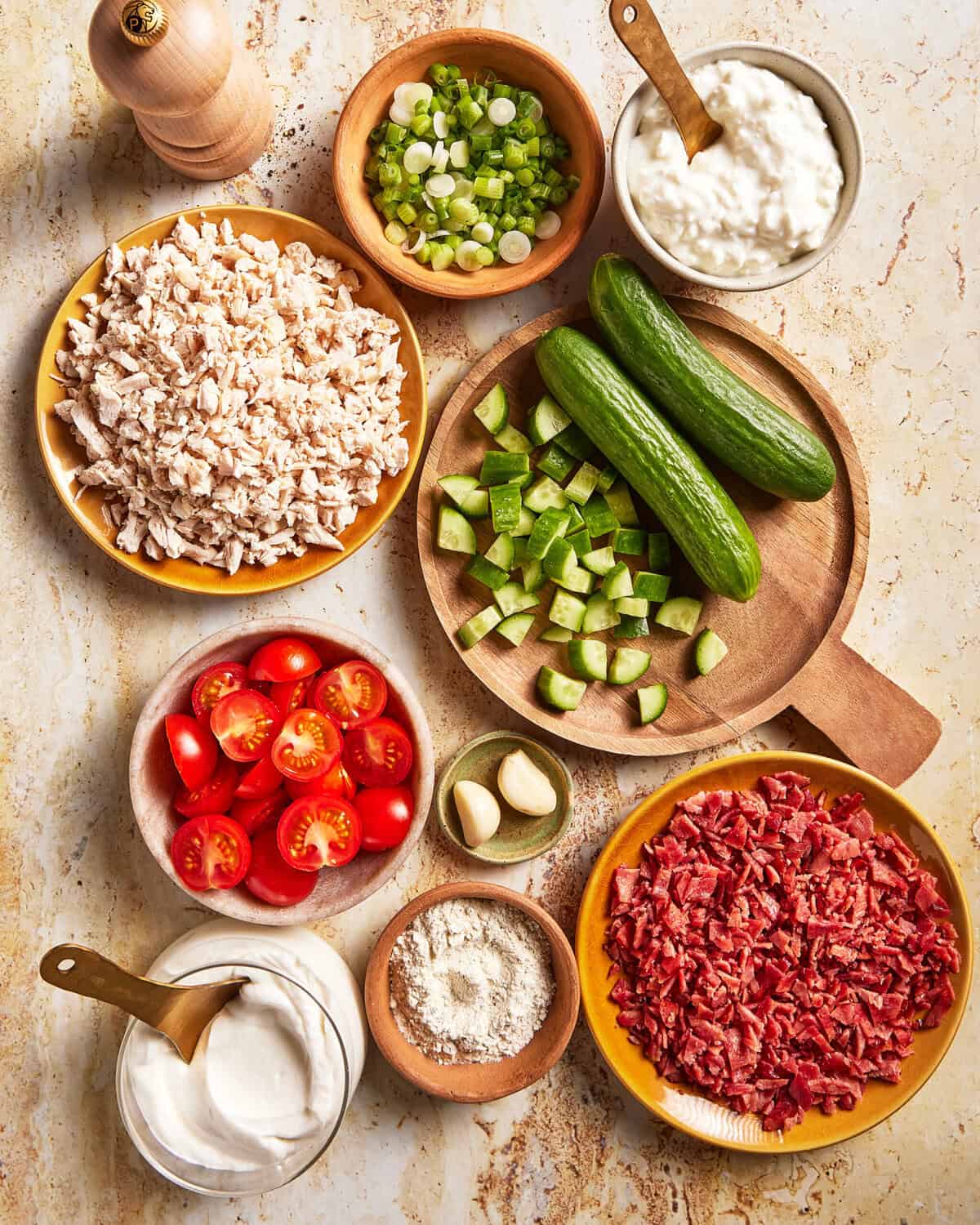 Bowls containing chopped cooked chicken, sliced green onions, cottage cheese, cucumbers, grape tomatoes, garlic cloves, sour cream, flour, and cooked bacon pieces arranged on a marble surface.
