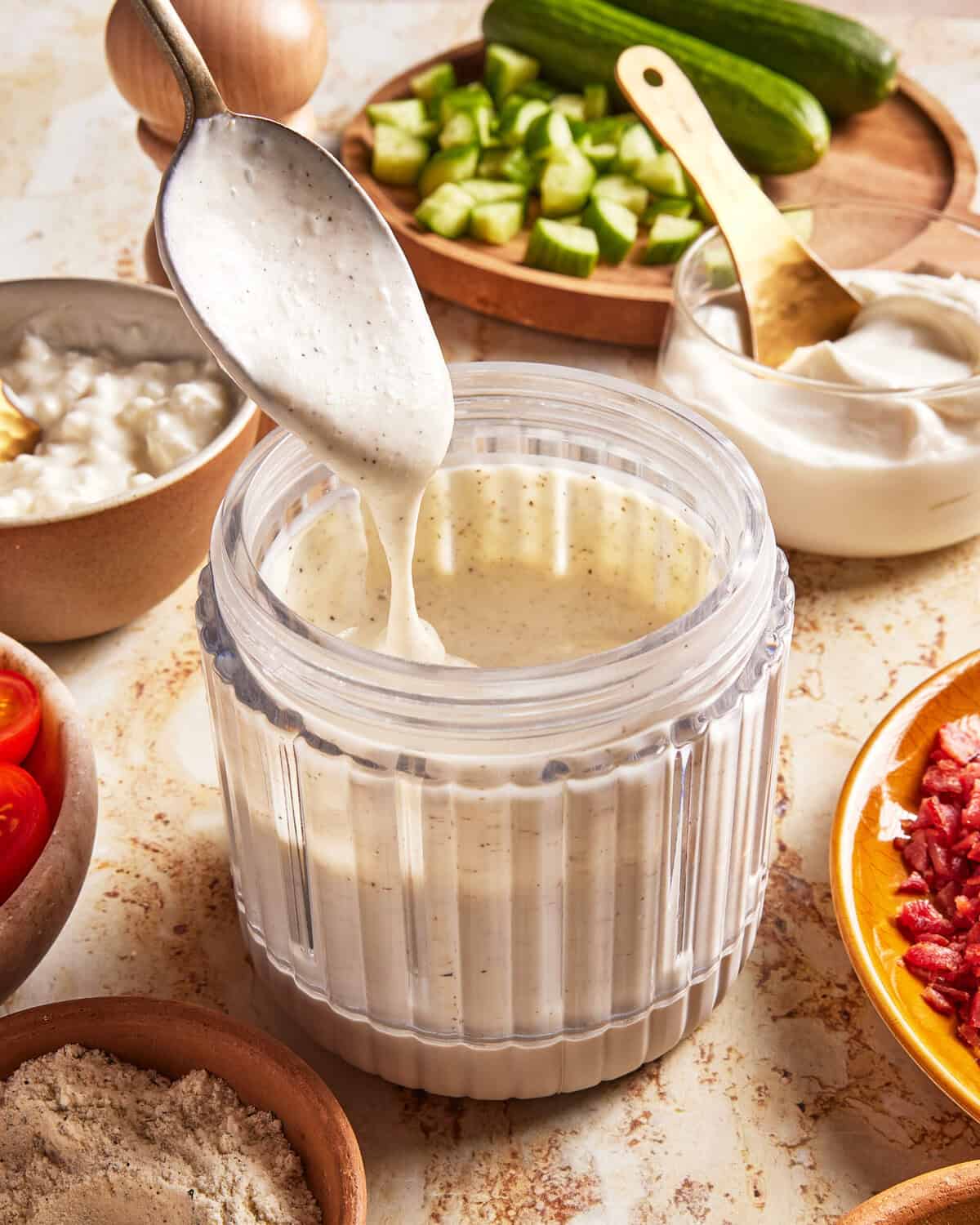 A spoon drizzles creamy white dressing from a jar, surrounded by chopped cucumbers, sour cream, tomatoes, bacon bits, and flour on a kitchen counter.