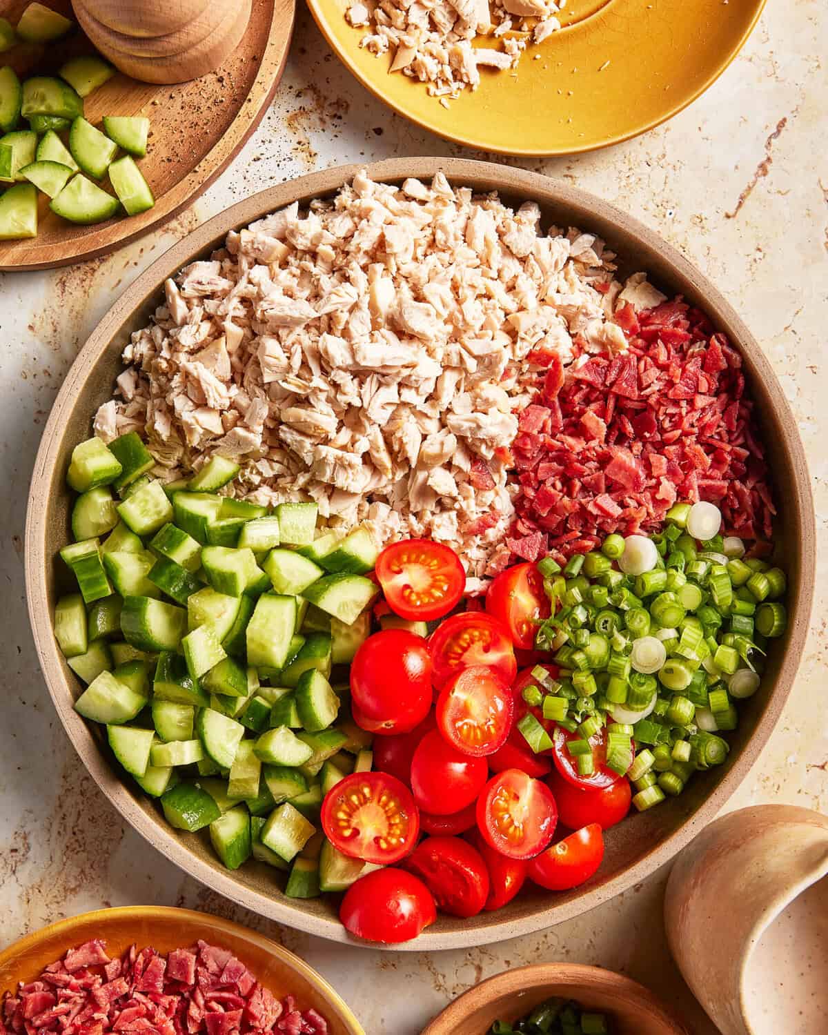 A bowl with sections of chopped chicken, bacon, cucumbers, grape tomatoes, and green onions. Surrounding plates hold more of the same ingredients, all on a light-colored surface.