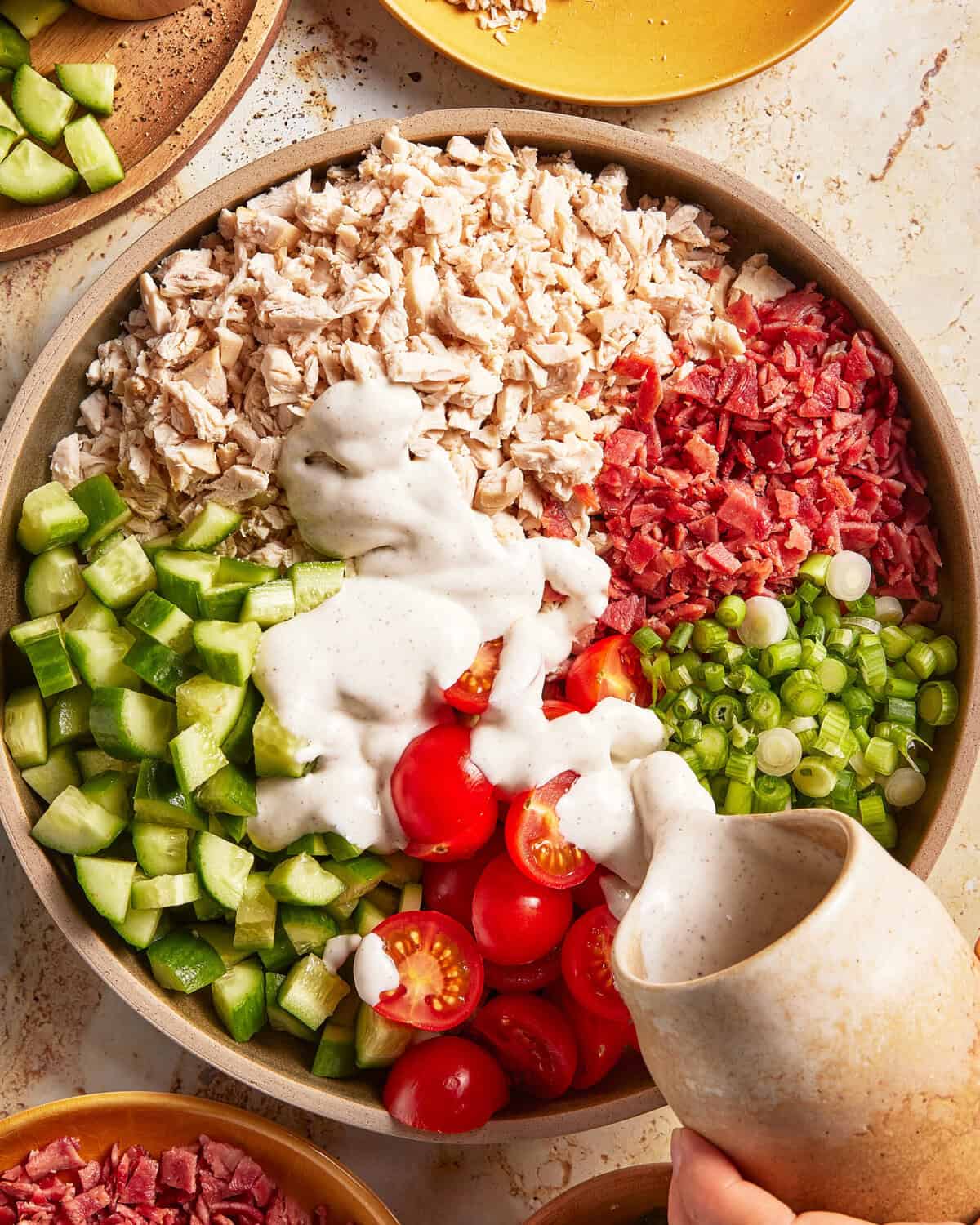 A large bowl with chopped chicken, bacon, cucumbers, green onions, cherry tomatoes, and ranch dressing being poured over the top.