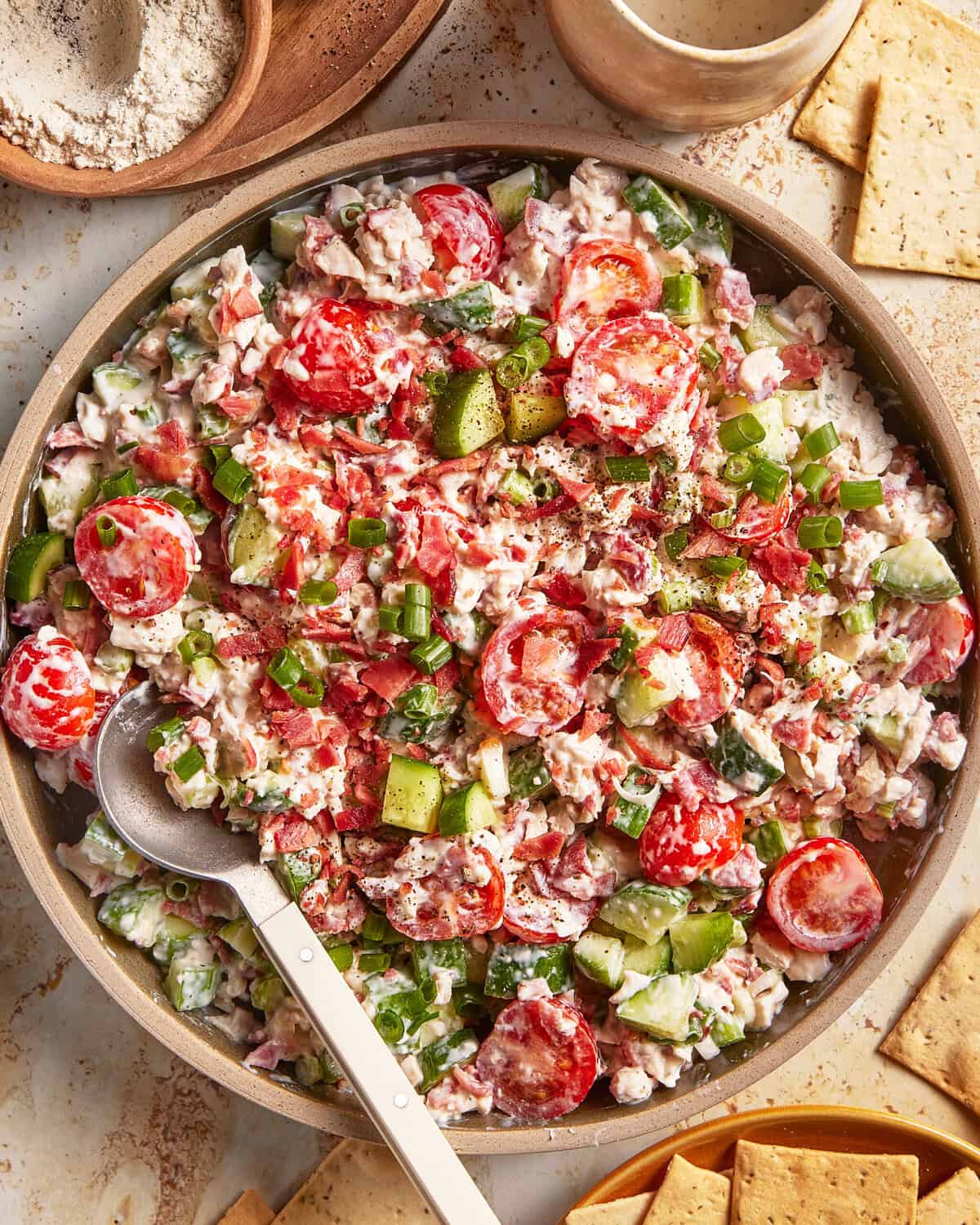 A bowl of creamy salad with cherry tomatoes, cucumbers, green onions, and herbs, with a serving spoon. Crackers and a small bowl of dressing are nearby.