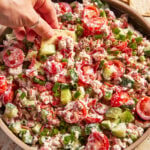 A hand dips a cracker into a bowl of creamy salad with cherry tomatoes, cucumbers, green onions, and a sprinkle of seasoning.