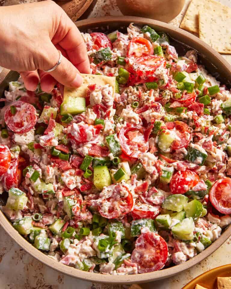 A hand dips a cracker into a bowl of creamy salad with cherry tomatoes, cucumbers, green onions, and a sprinkle of seasoning.
