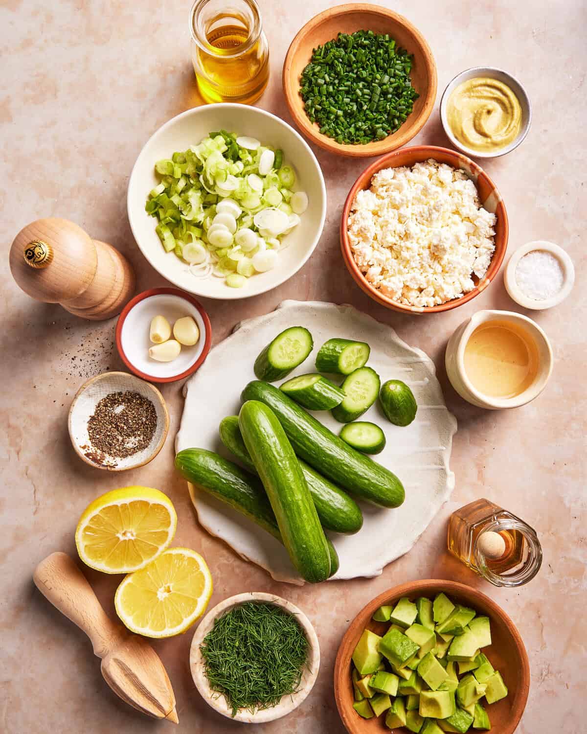 An overhead view of ingredients in bowls and on a plate, including cucumbers, chopped green onions, chives, feta cheese, garlic, lemon halves, avocado, dill, olive oil, vinegar, mustard, salt, and pepper.