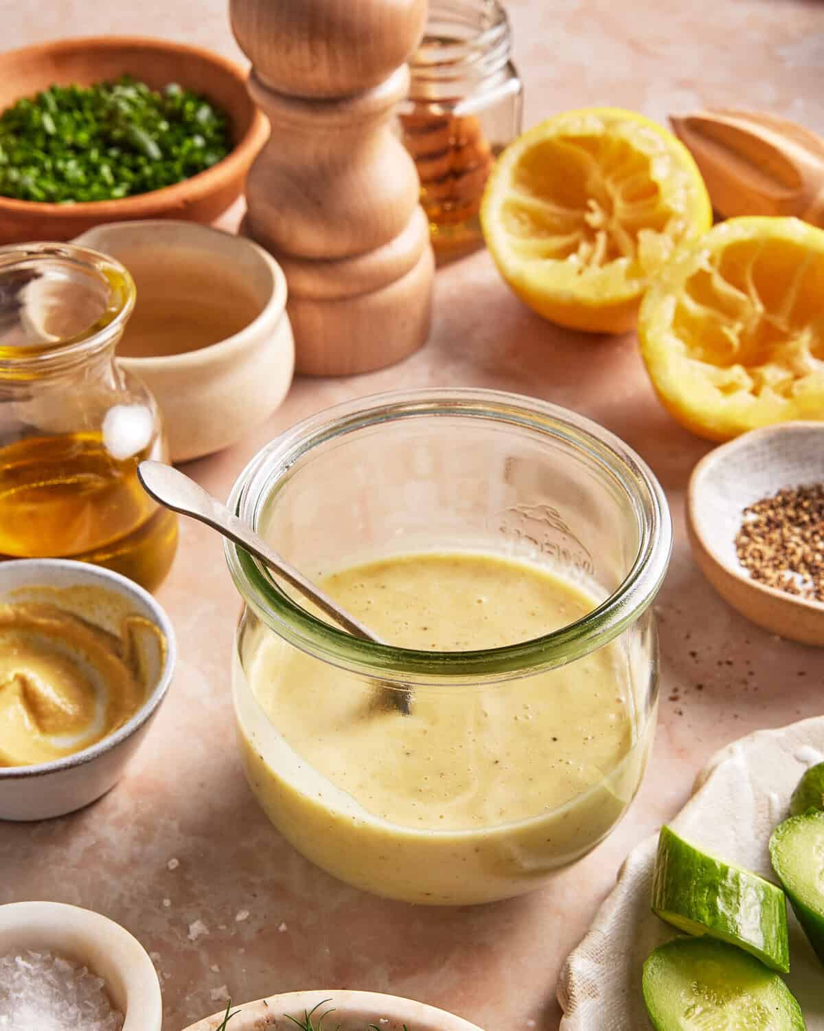 A jar of creamy dressing with a spoon sits on a counter surrounded by ingredients: mustard, olive oil, chives, ground pepper, sea salt, honey, cucumber slices, and halved lemons.