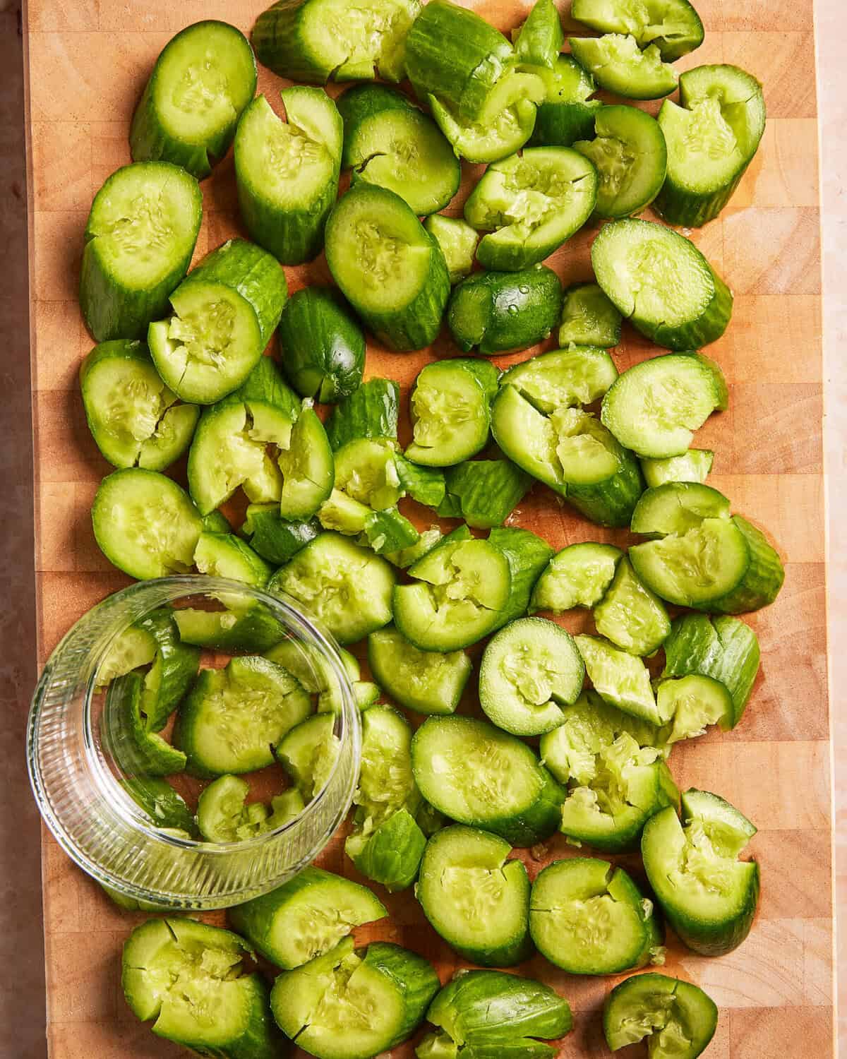 Smashed pieces of cucumber scattered across a wooden cutting board, with a glass tumbler lying on its side, suggesting it was used to smash the cucumbers.
