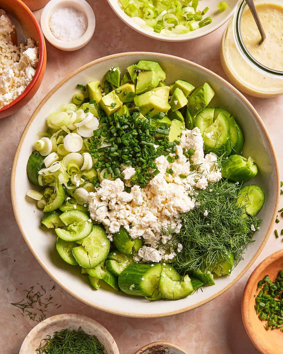 A large bowl filled with chopped cucumber, avocado, scallions, crumbled feta cheese, fresh dill, and chives, surrounded by small bowls of salt, feta, herbs, and a jar of salad dressing.
